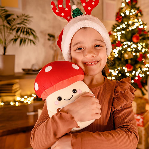 Child wearing a festive hat with reindeer antlers, holding a plush toy, in front of a decorated Christmas tree.