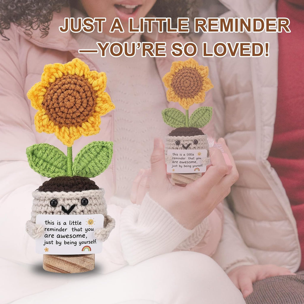 Person holding a crocheted sunflower plant with a message card, surrounded by text 'JUST A LITTLE REMINDER — YOU'RE SO LOVED!'.